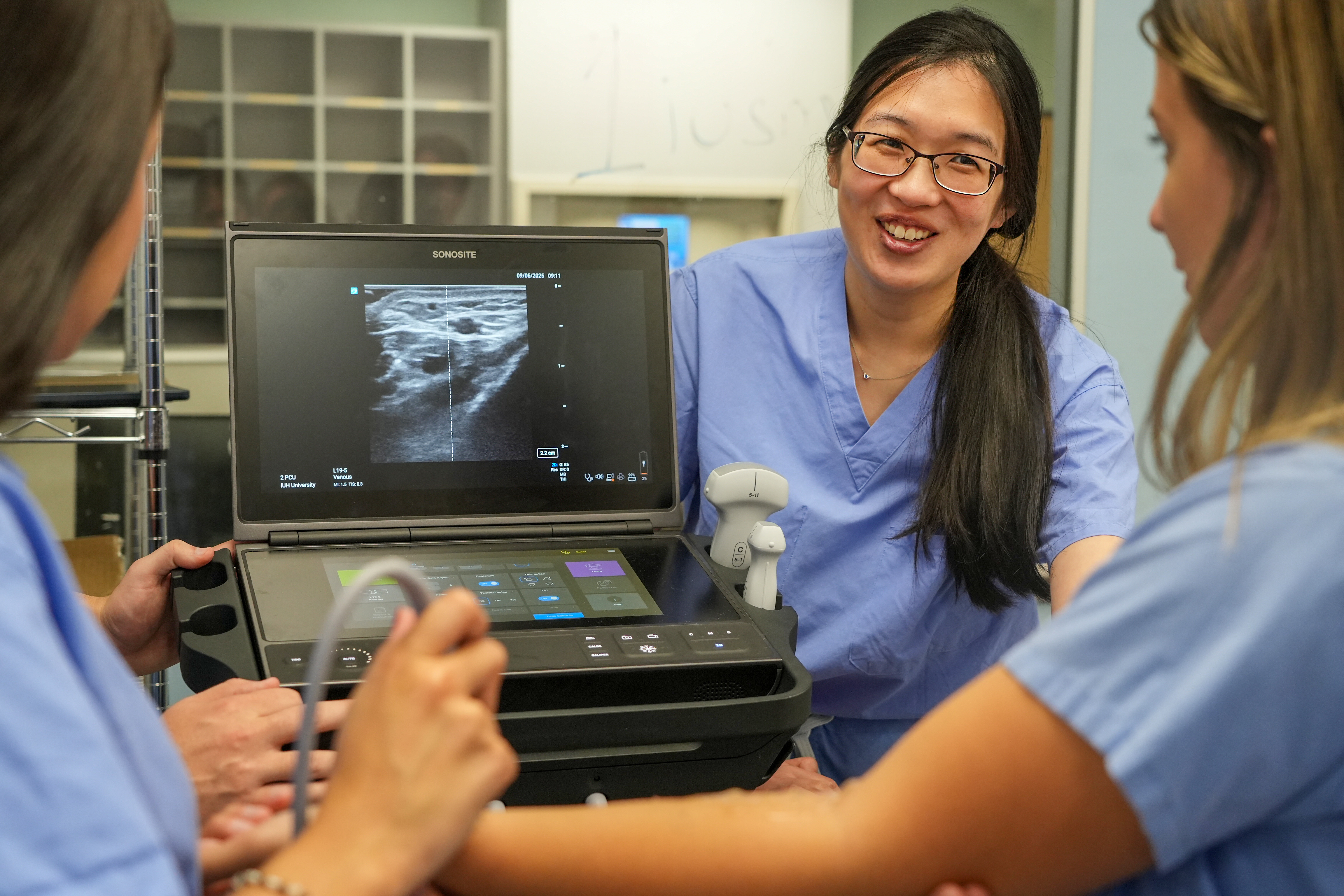 Clinician demonstrates a portable ultrasound machine to colleagues, with an imaging display visible on the screen.