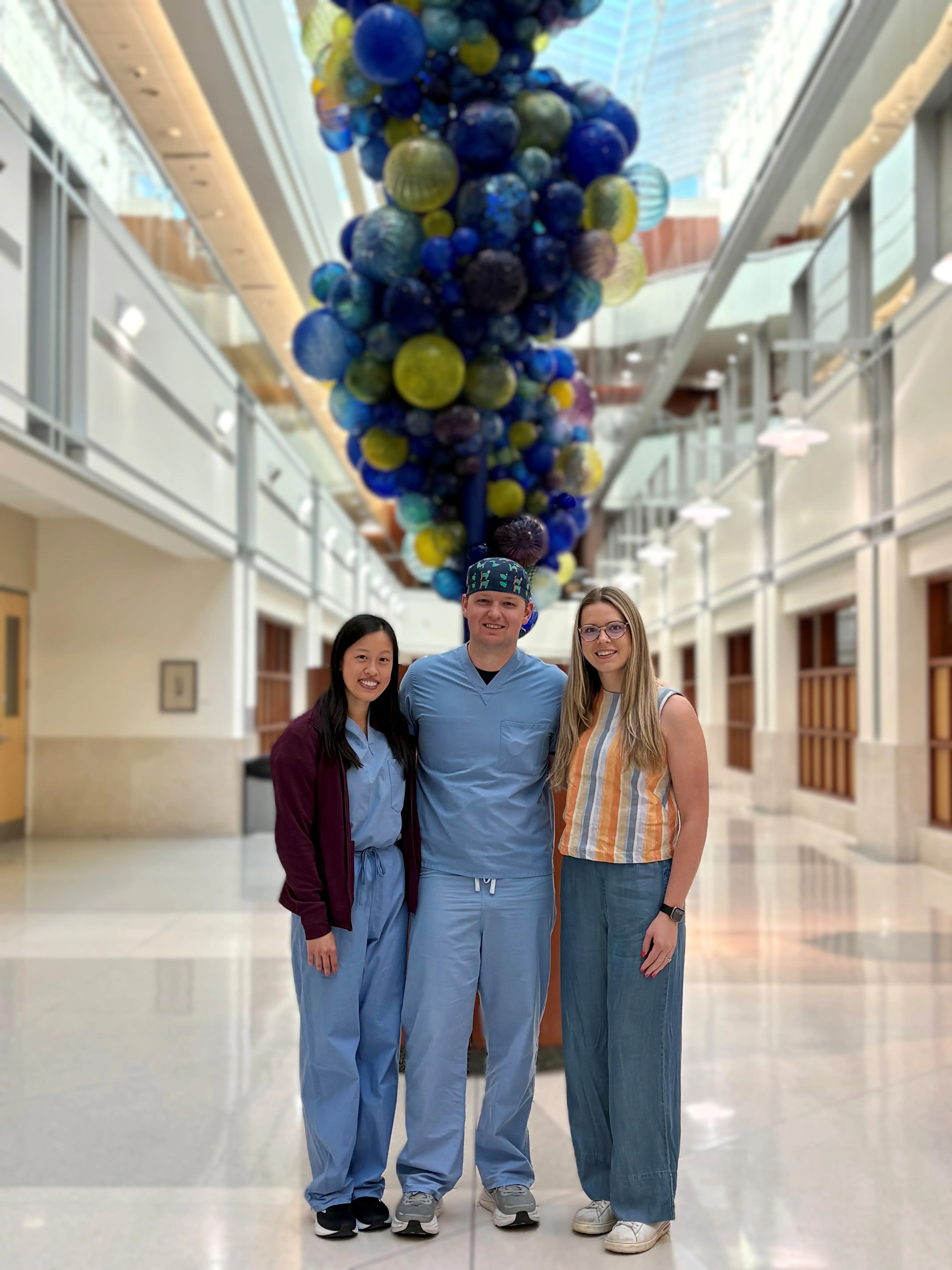Three people pose in a bright hospital atrium beneath a large hanging sculpture of blue and green glass spheres.