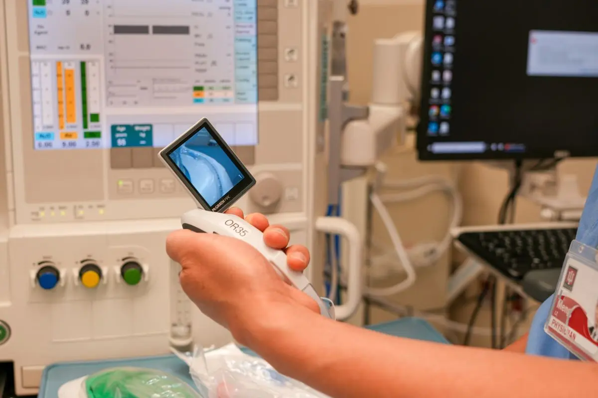 Close-up of a clinician’s hand holding a handheld medical scope displaying an internal airway image, with monitoring equipment in the background.