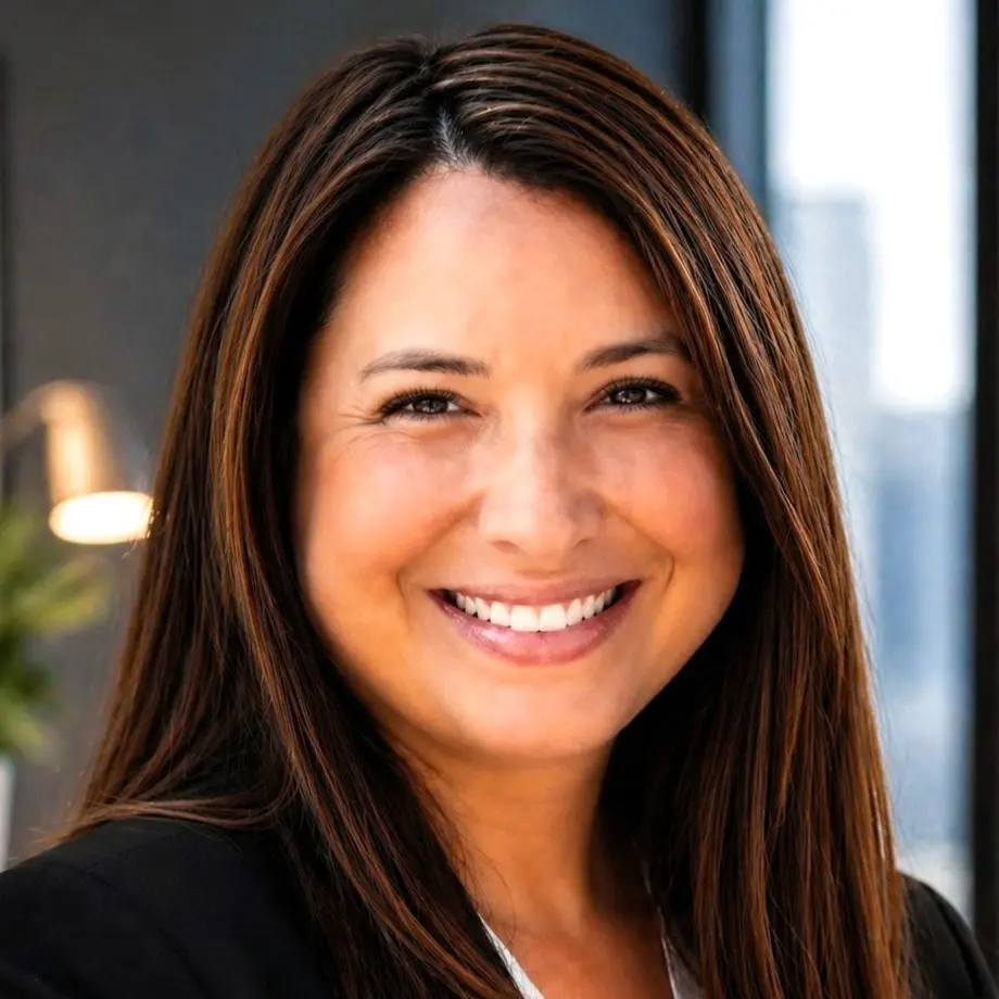 Professional headshot of a person in a black blazer and white top in an office setting.