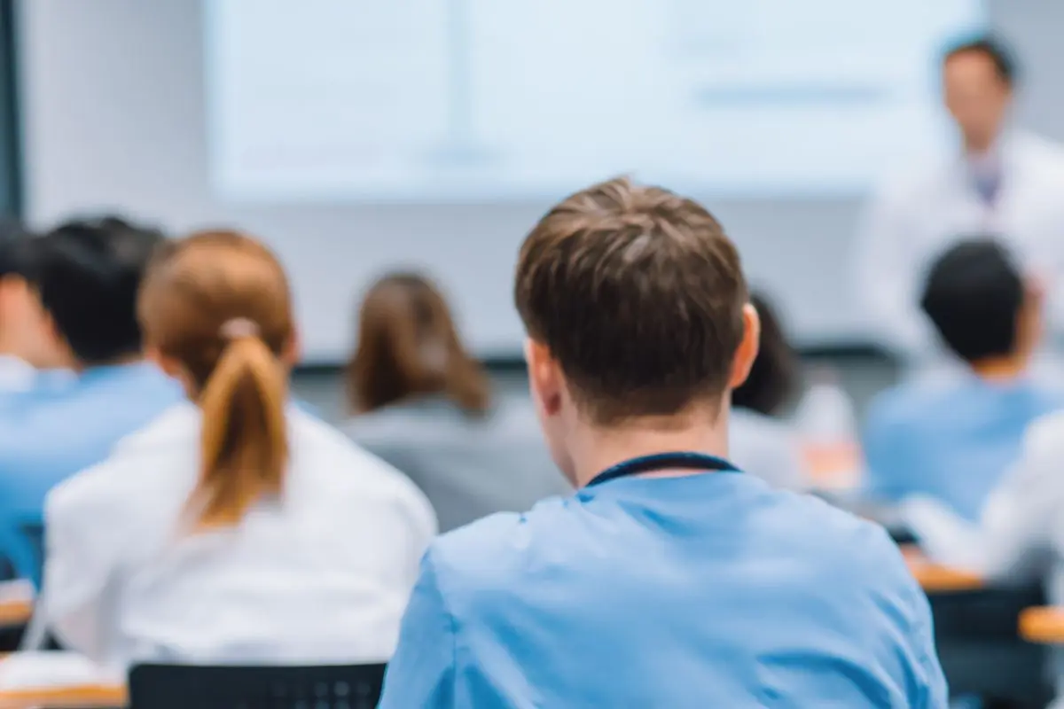 Group of people in a classroom facing forward while a presenter speaks at the front, with the foreground showing the back of a person in blue scrubs.