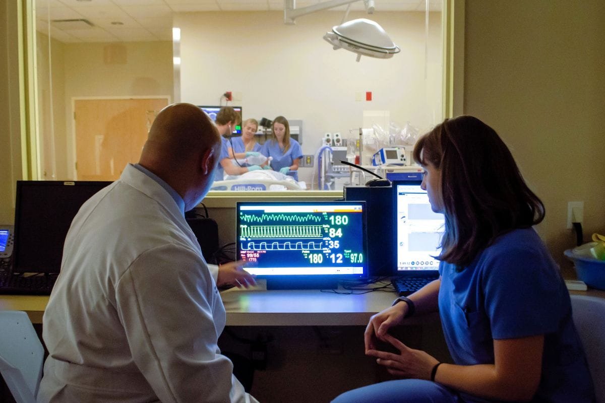 Clinicians review vital signs on a monitor while a team treats a patient in a simulation room behind glass.