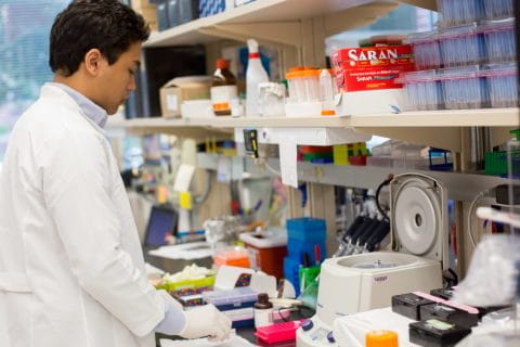 A researcher in a white coat and gloves prepares samples in the lab.