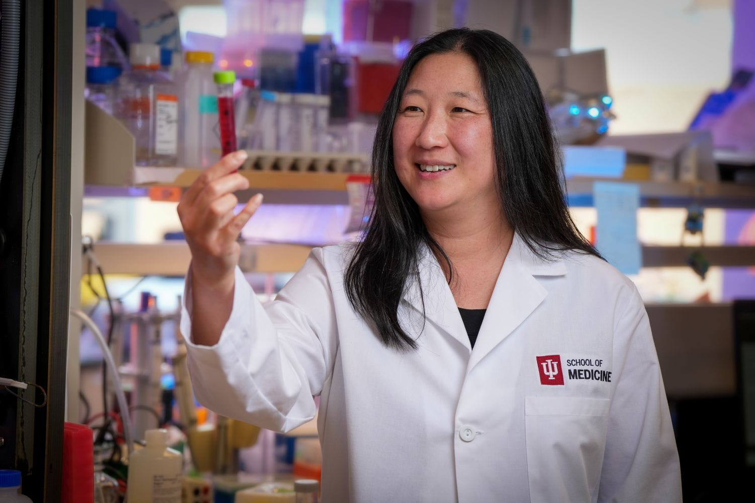Elizabeth Yeh holds a lab sample up to eye level. She's wearing a white lab coat with shelves of equipment and tools on the lab bench behind her.