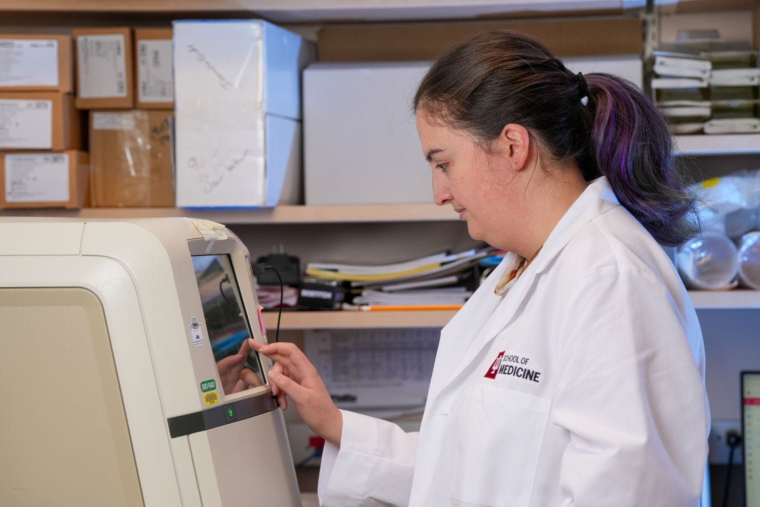 A student wearing a lab coat with her hair pulled back keys in instructions on a lab instrument.