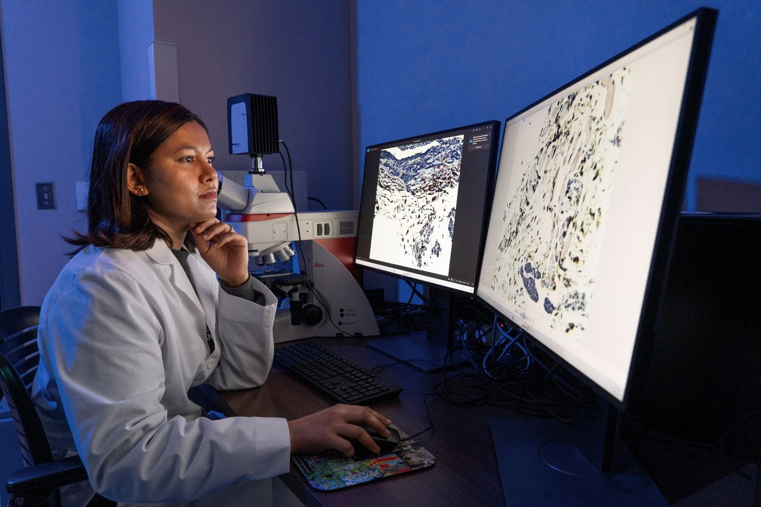A researcher reviews microscopy scans on two bright monitors in a dark room.