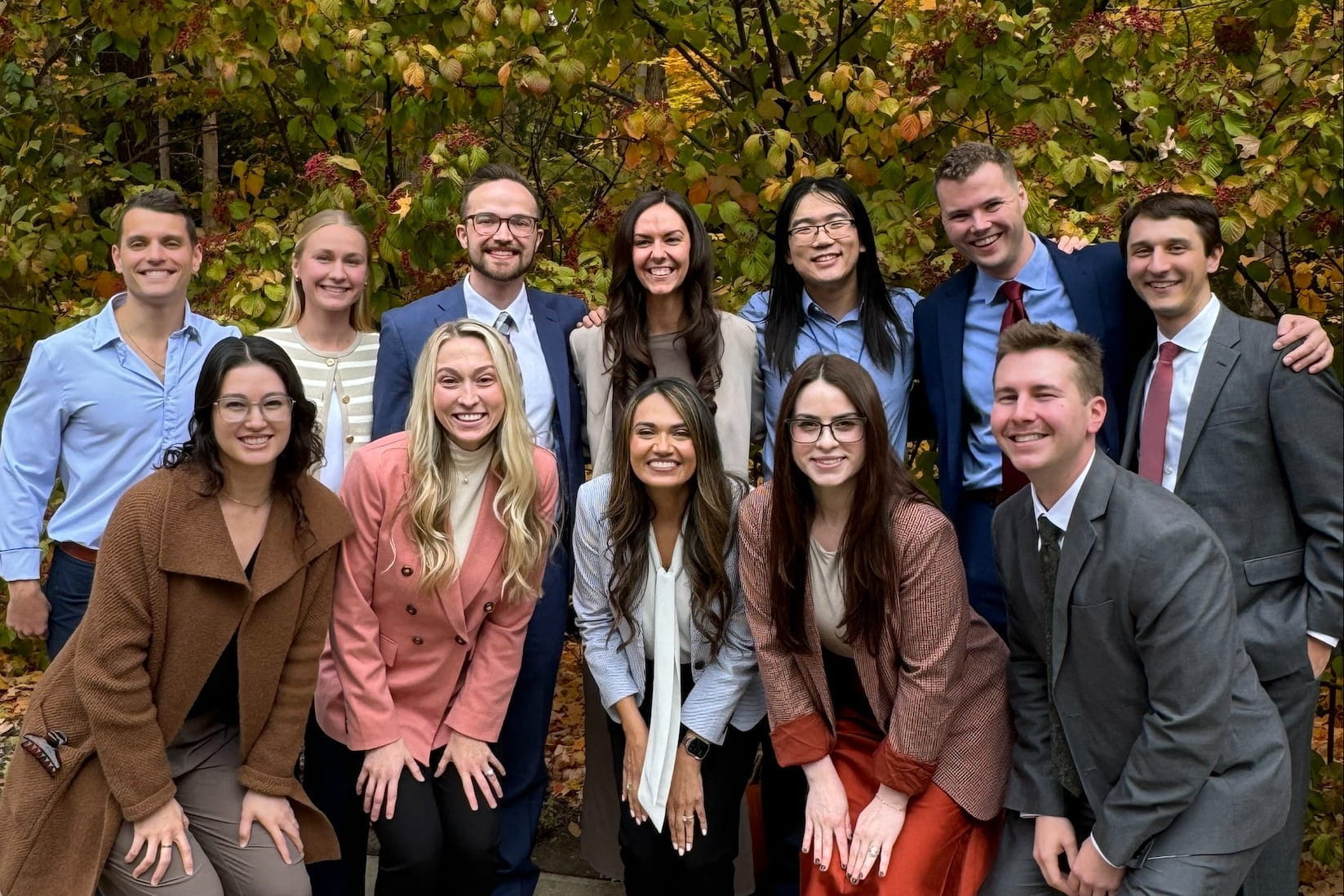 12 residents pose for a group photo on a sunny day. Shrubs in fall colors fill the background behind the group.