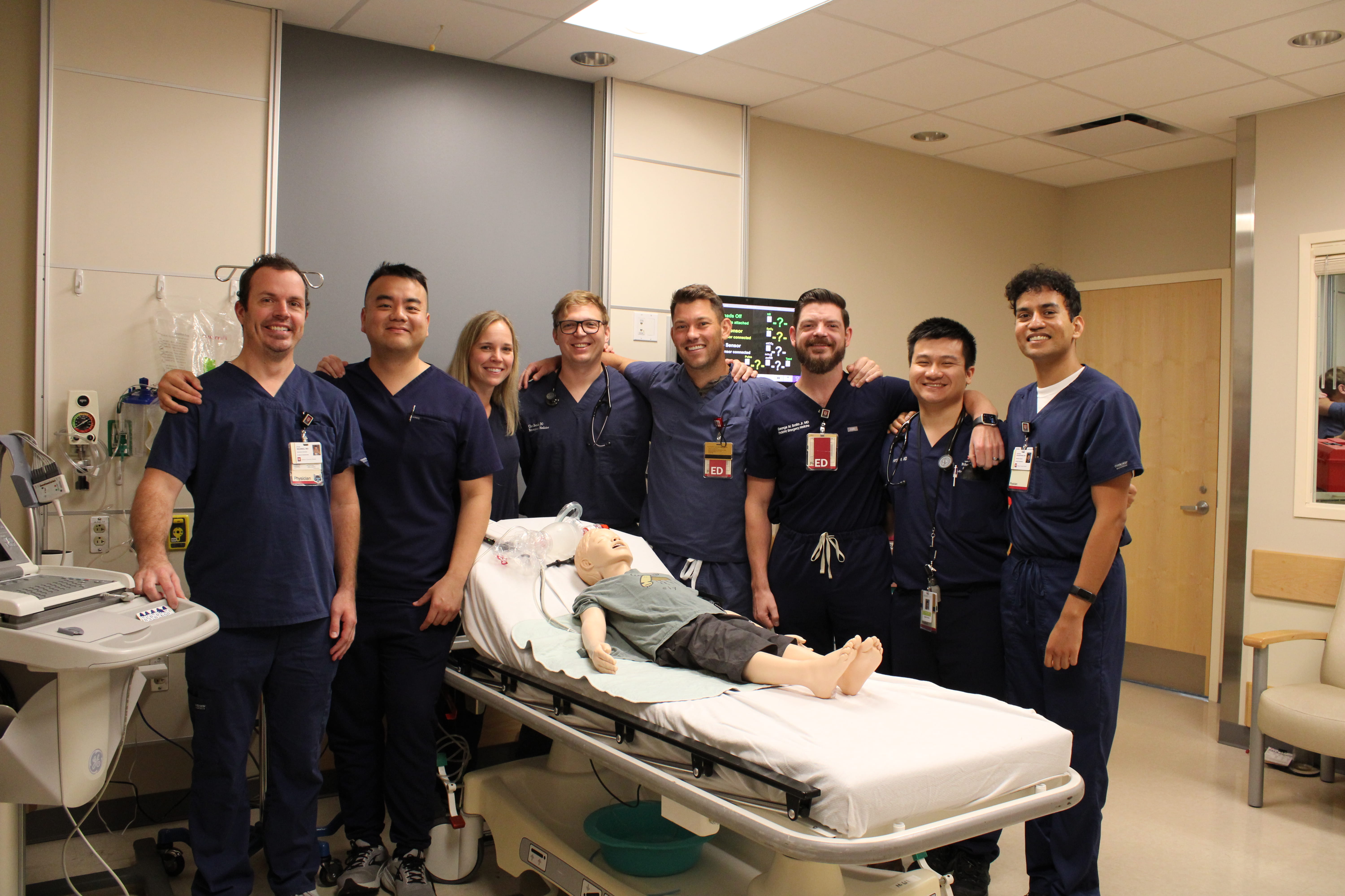 A group of PEM fellows smiling at the camera and surrounding a pediatric patient mannequin.