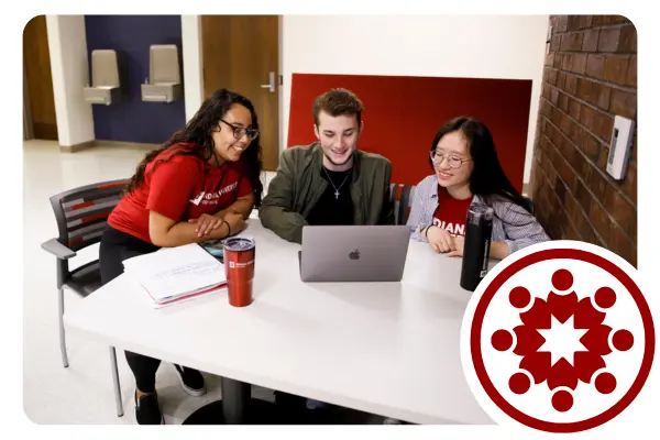 Three college students sit together at a table while working. One student works on a laptop while the others lean in collaboratively.