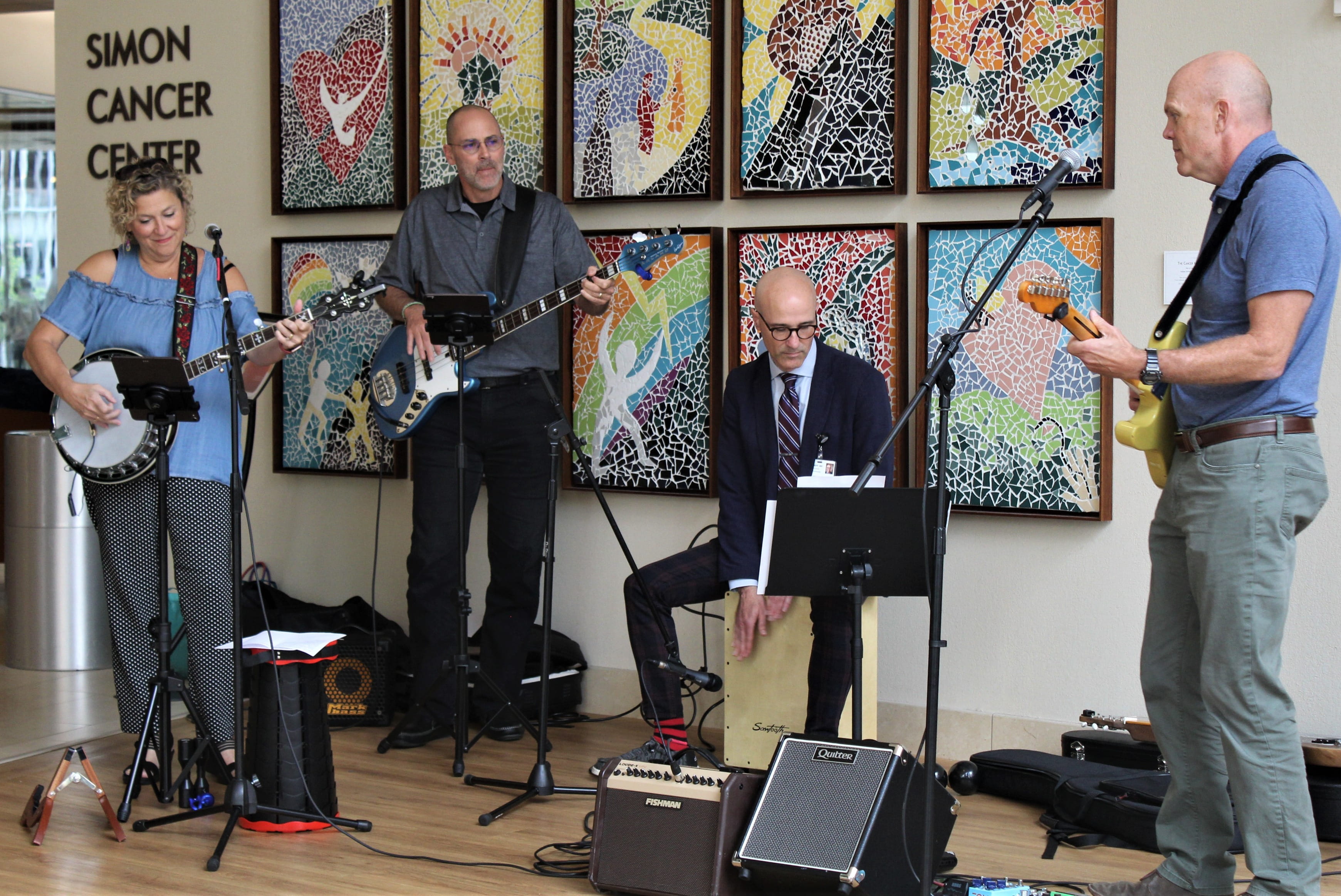 Department of Medicine Chair David Aronoff sits atop a wooden, box-like cajon drum in the lobby of the Simon Cancer Center as he plays with three other members of the band "the Crooked Finger Rhythm Review." The band includes a banjo player and two guitar players. Colorful artwork is displayed on the wall behind the band. 