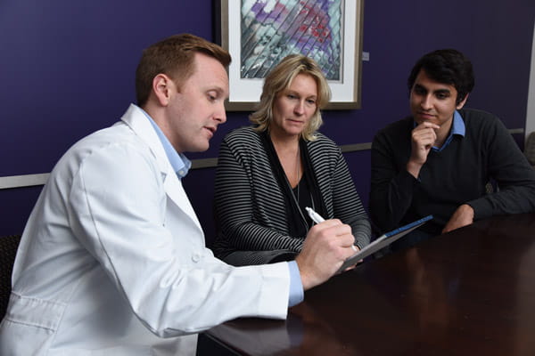 A physician in a white coat and two researchers talk together and review notes on a notepad