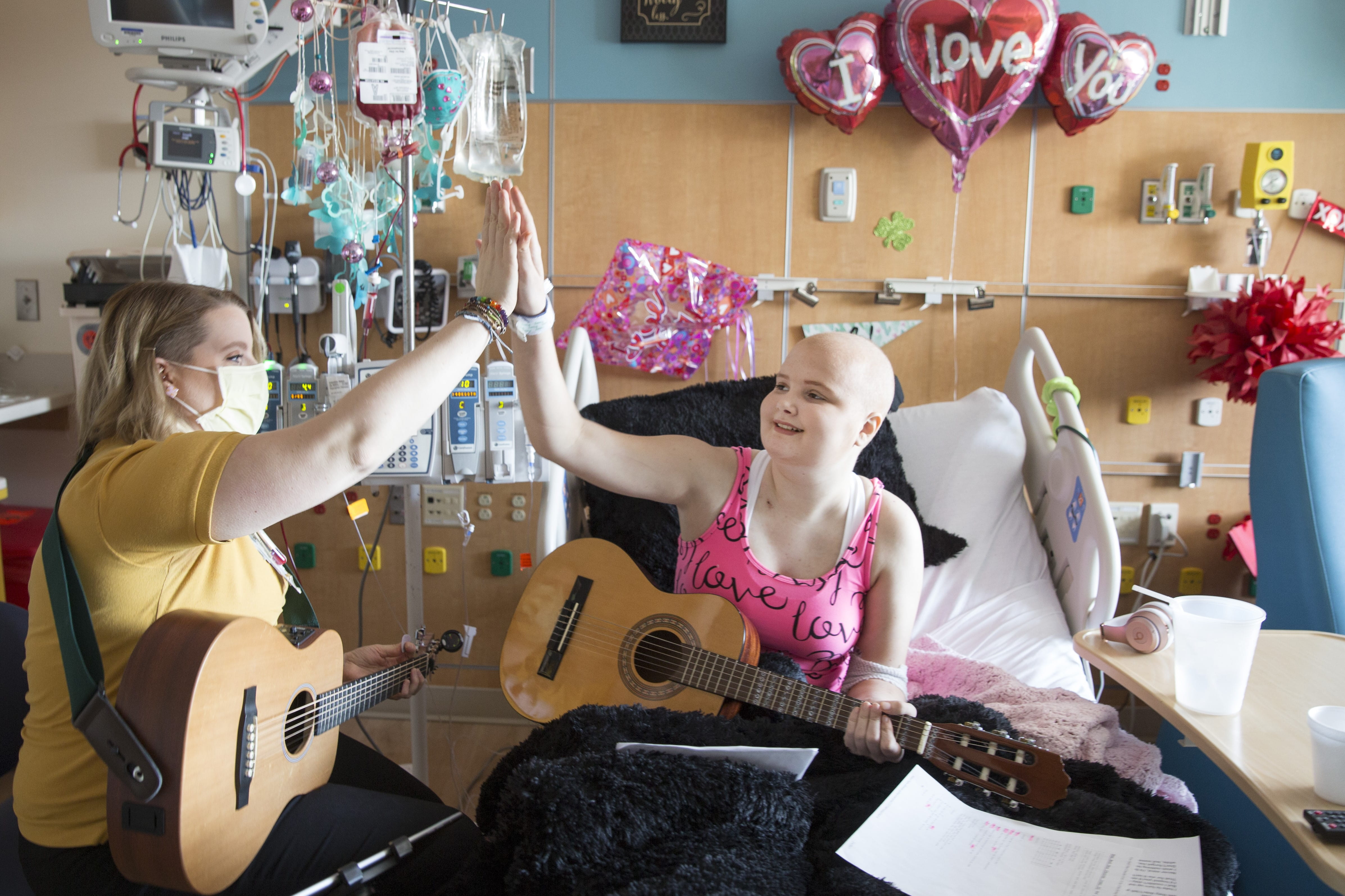 A teen girl with a bald head holds a guitar while she sits in a hospital bed surrounded by valentine's day balloons. She is high fiving a music therapist who also holds a guitar.