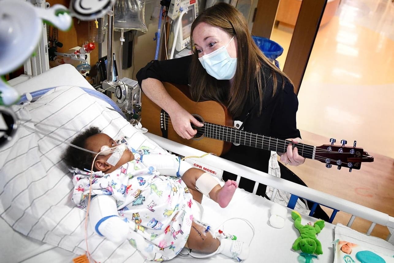 a music therapist at Riley Hospital sings and plays guitar for a baby in a hospital bed.