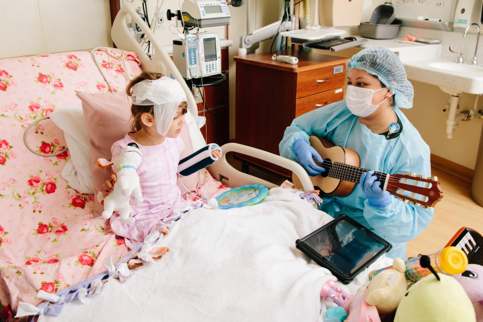 A music therapist in scrubs and mask plays the guitar. A young patient with her head wrapped in bandages sits in a hospital bed and sings along while holding a stuffed toy dog.