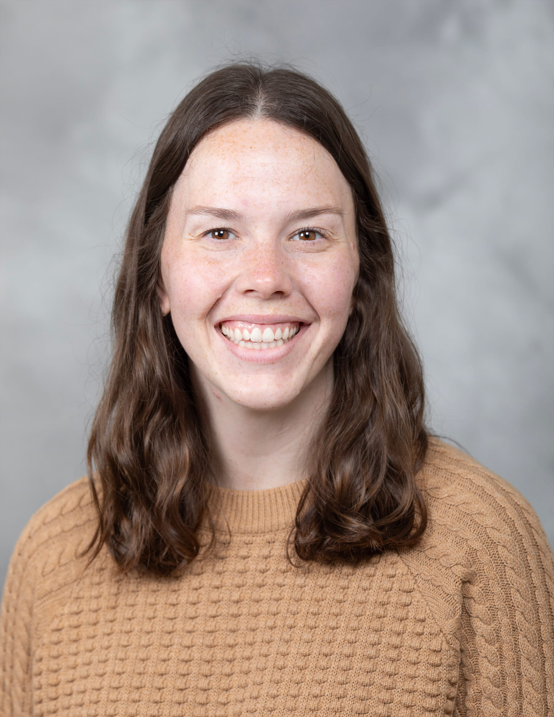 Woman with brown, curly hair is wearing a tan sweater and smiling.