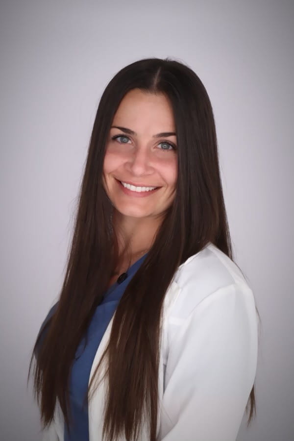 Woman with long, straight, brown hair wearing a white lab coat and blue shirt is smiling. 