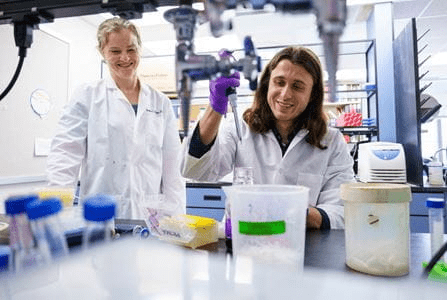 A laboratory scene with two people in white lab coats working at a bench surrounded by scientific equipment and containers.
