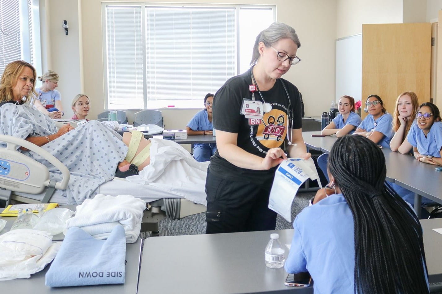 A nurse educator shows a resident intern the blood pressure cuff. Other interns watch from the side. A simulated patient sits on the hospital bed in the classroom.