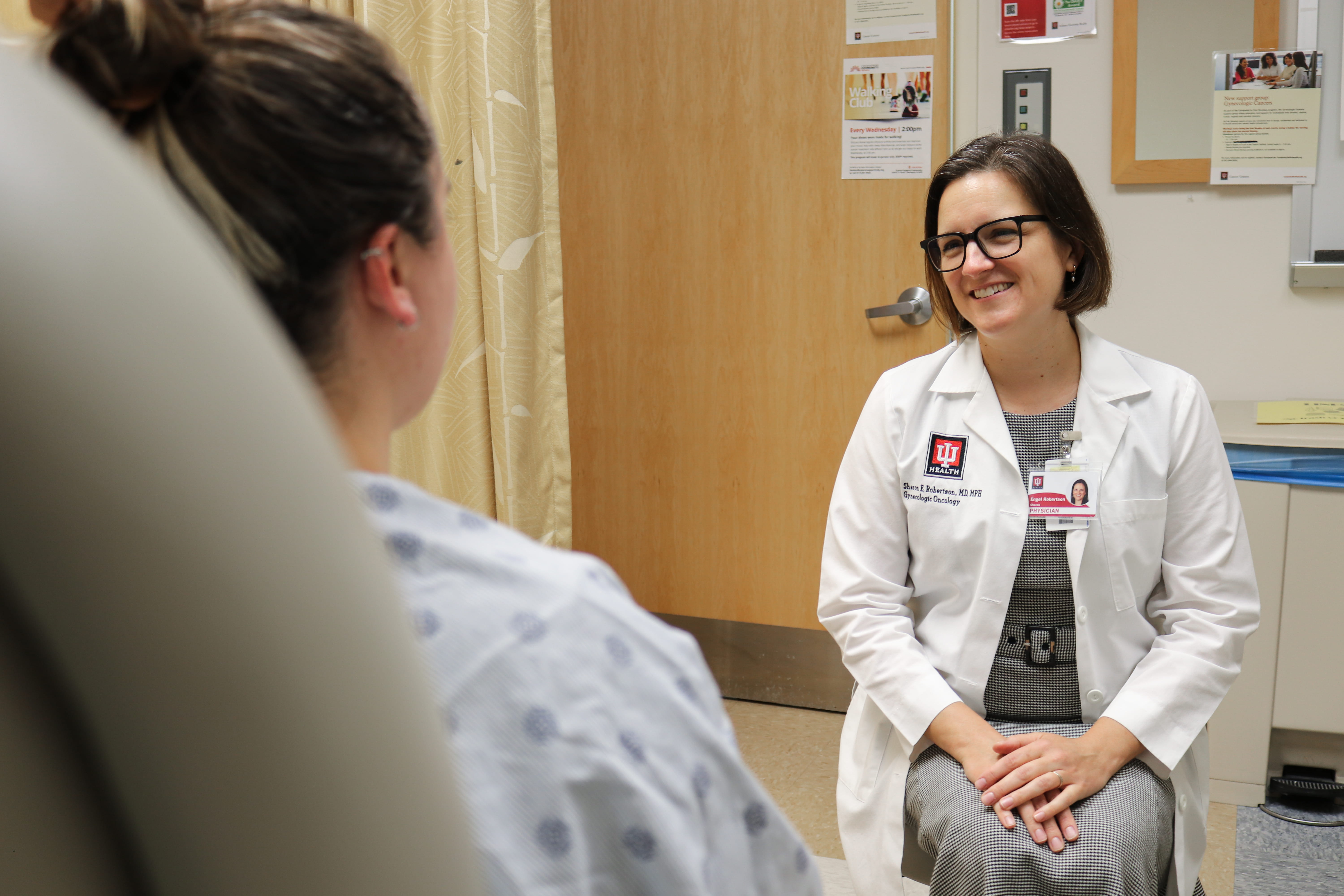Sharon Robertson, MD, wears her white lab coat while sitting in front of a patient. The patient is seated in a chair in the clinic with her back to the camera. Robertson smiles while listening to the patient.