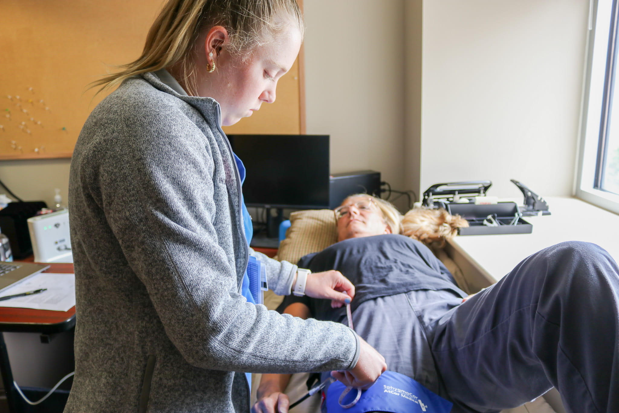 A third-year medical student performs a test on a standardized patient using a thigh cuff to obtain readings.