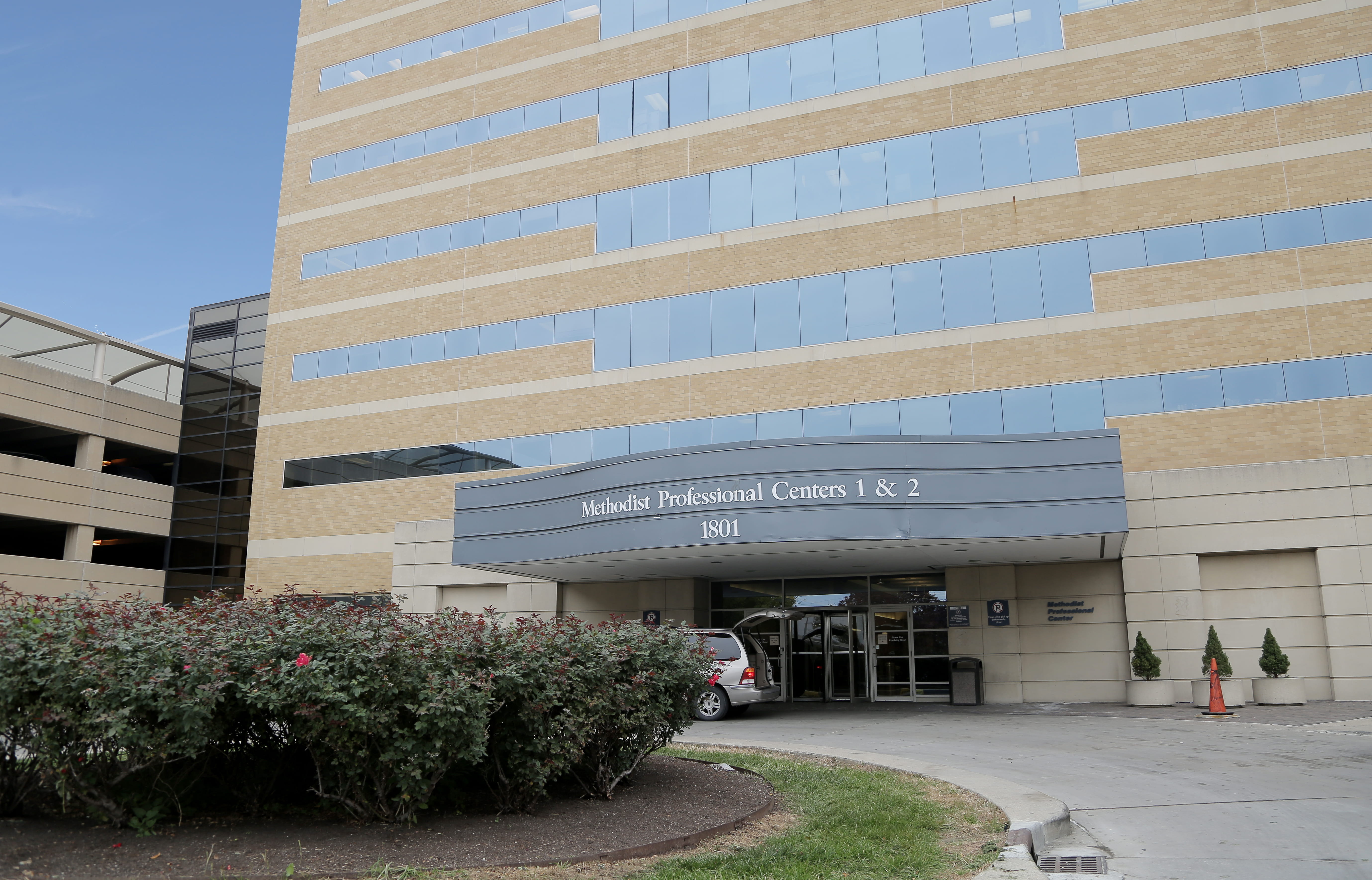 The front of a brick building with many rows of windows faces a bush in the foreground. An outcropping on the building has text reading: Methodist Professional Centers 1 & 2 1801. To the left of the building is a cropped parking garage and blue sky.