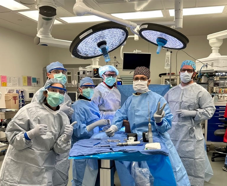 Several medical residents and physicians stand around an operating room table holding surgery tools. Each individual is wearing a surgery gown, gloves, caps, and glasses.