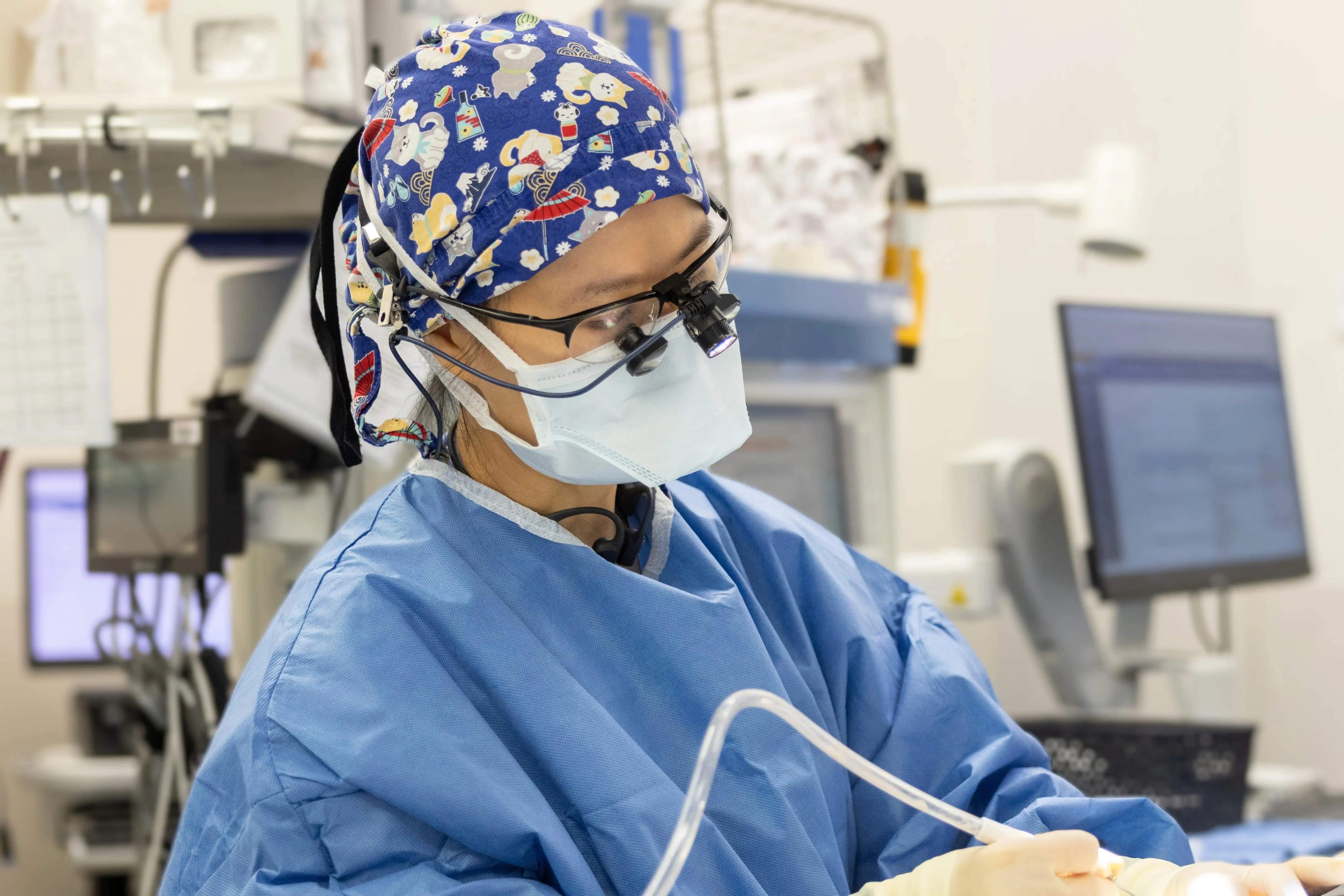 Clinician wearing surgical attire, mask, and magnifying loupes performs a procedure in an operating room.