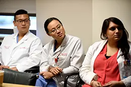 Three medical trainees in white coats seated in a small room, listening attentively, one holding a pen and taking notes.