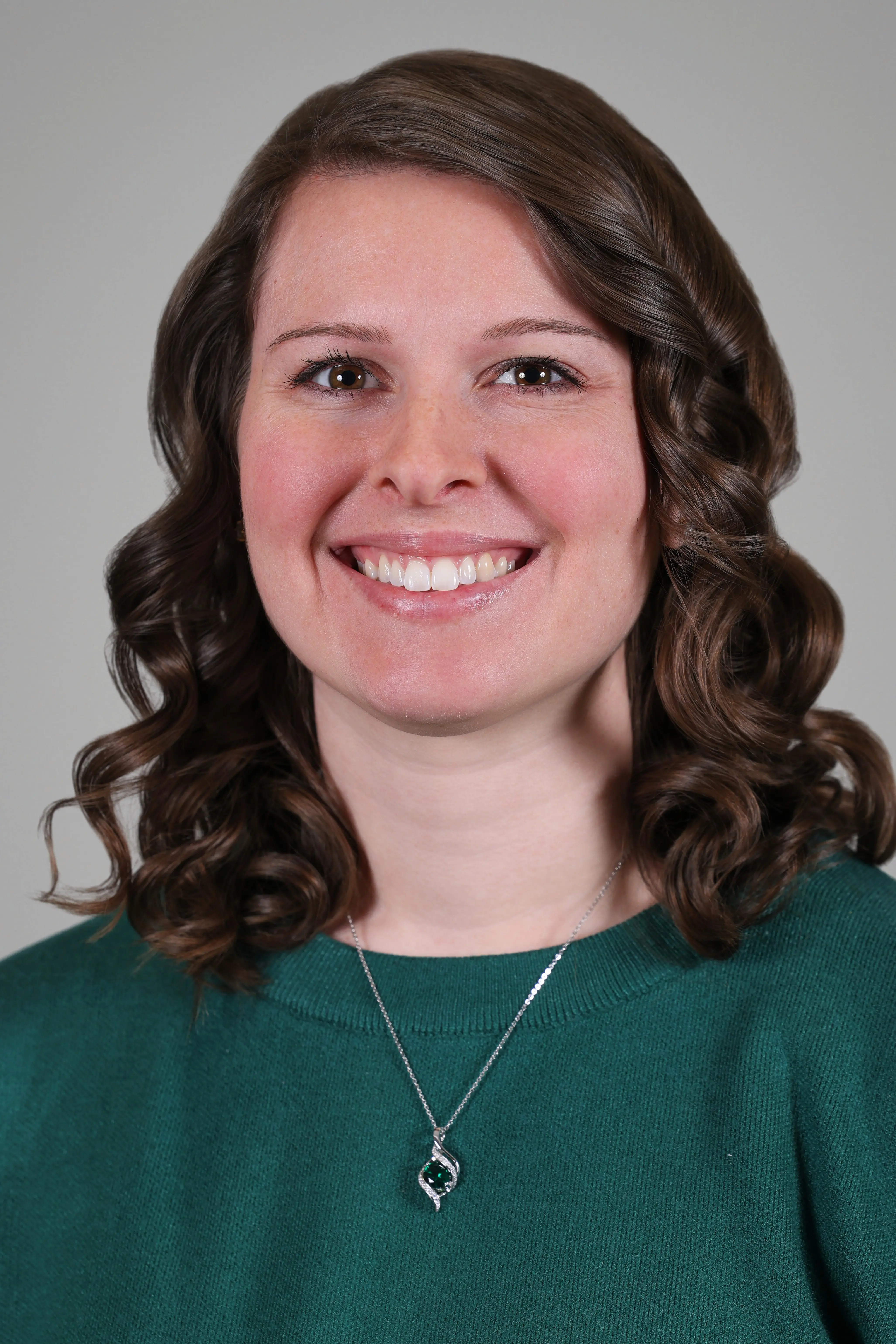 Headshot of a person wearing a green sweater and pendant necklace against a gray background.