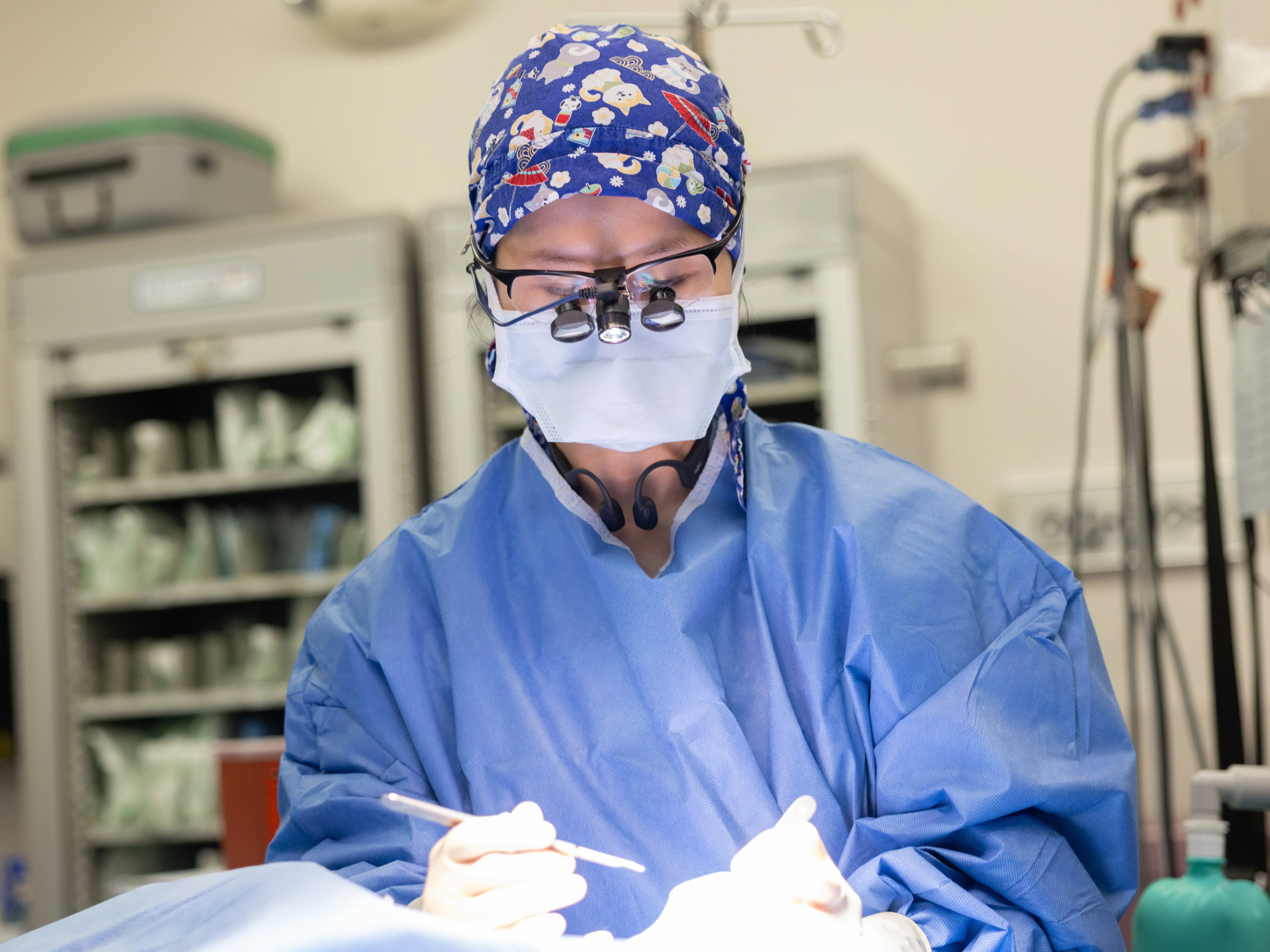 Masked surgeon wearing magnifying loupes and a surgical gown performs a procedure in an operating room surrounded by medical equipment.