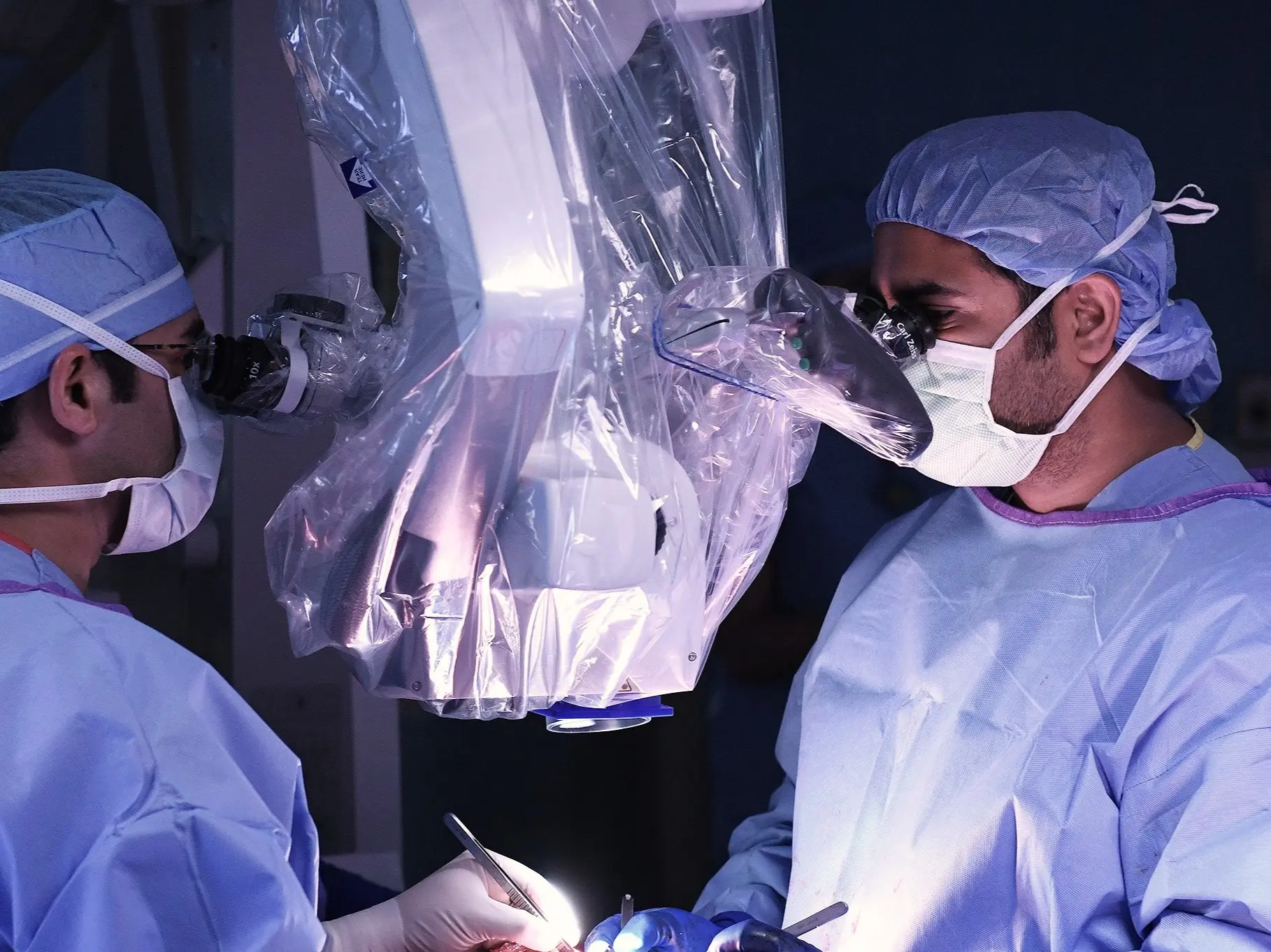 Two surgeons in sterile gowns, masks, and caps perform a procedure using an operating microscope in a surgical suite.