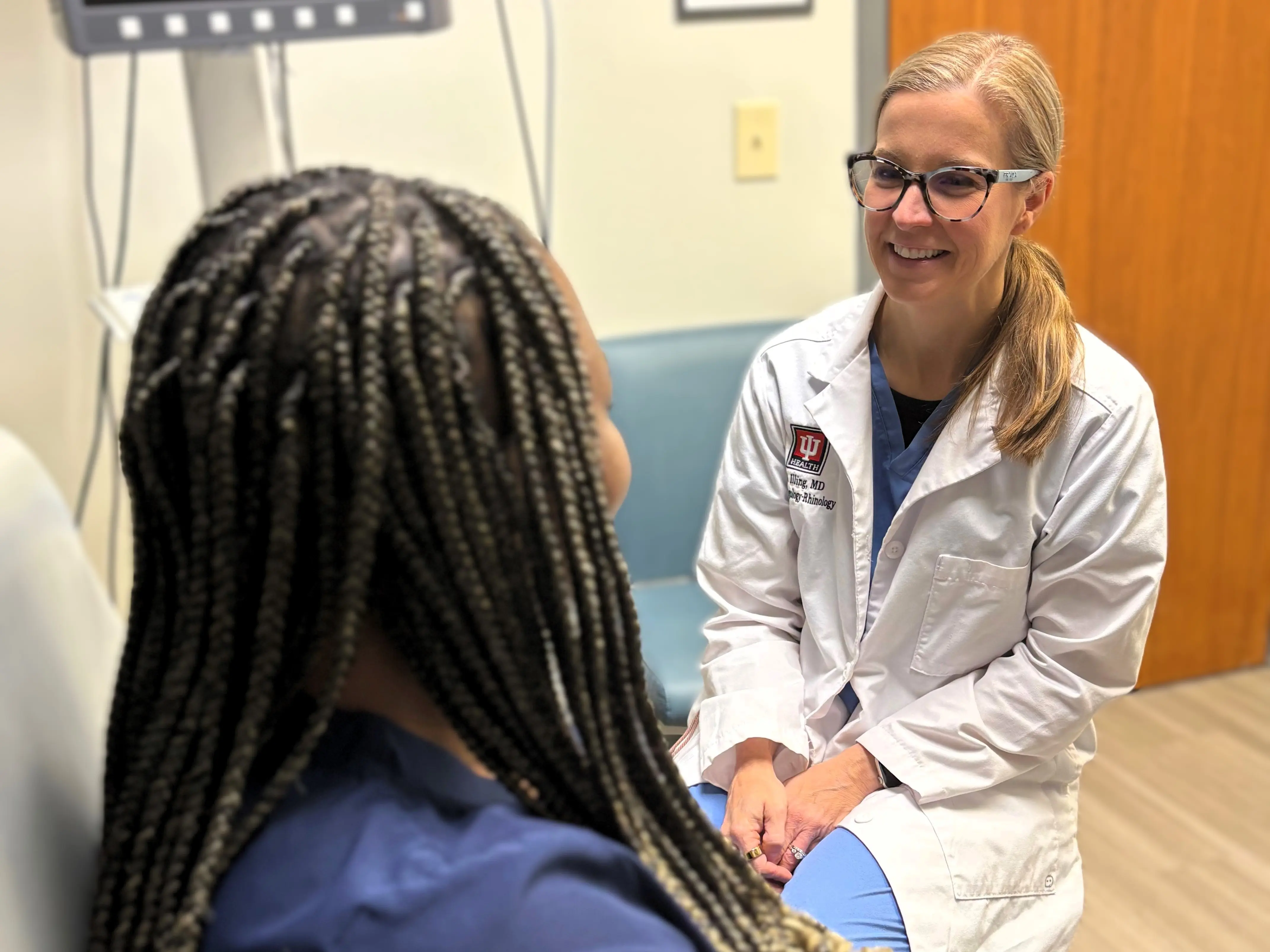 Clinician in a white coat seated in an exam room speaking with a patient during a consultation.