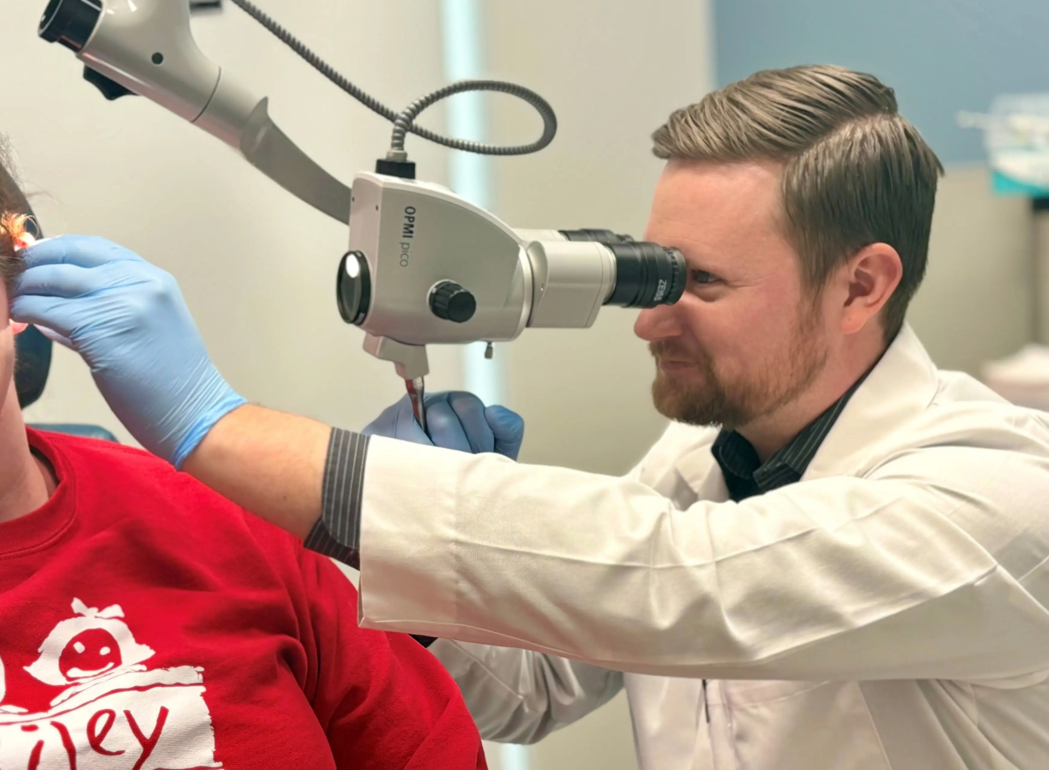 Clinician wearing gloves examines a patient’s ear using a surgical microscope during an office visit
