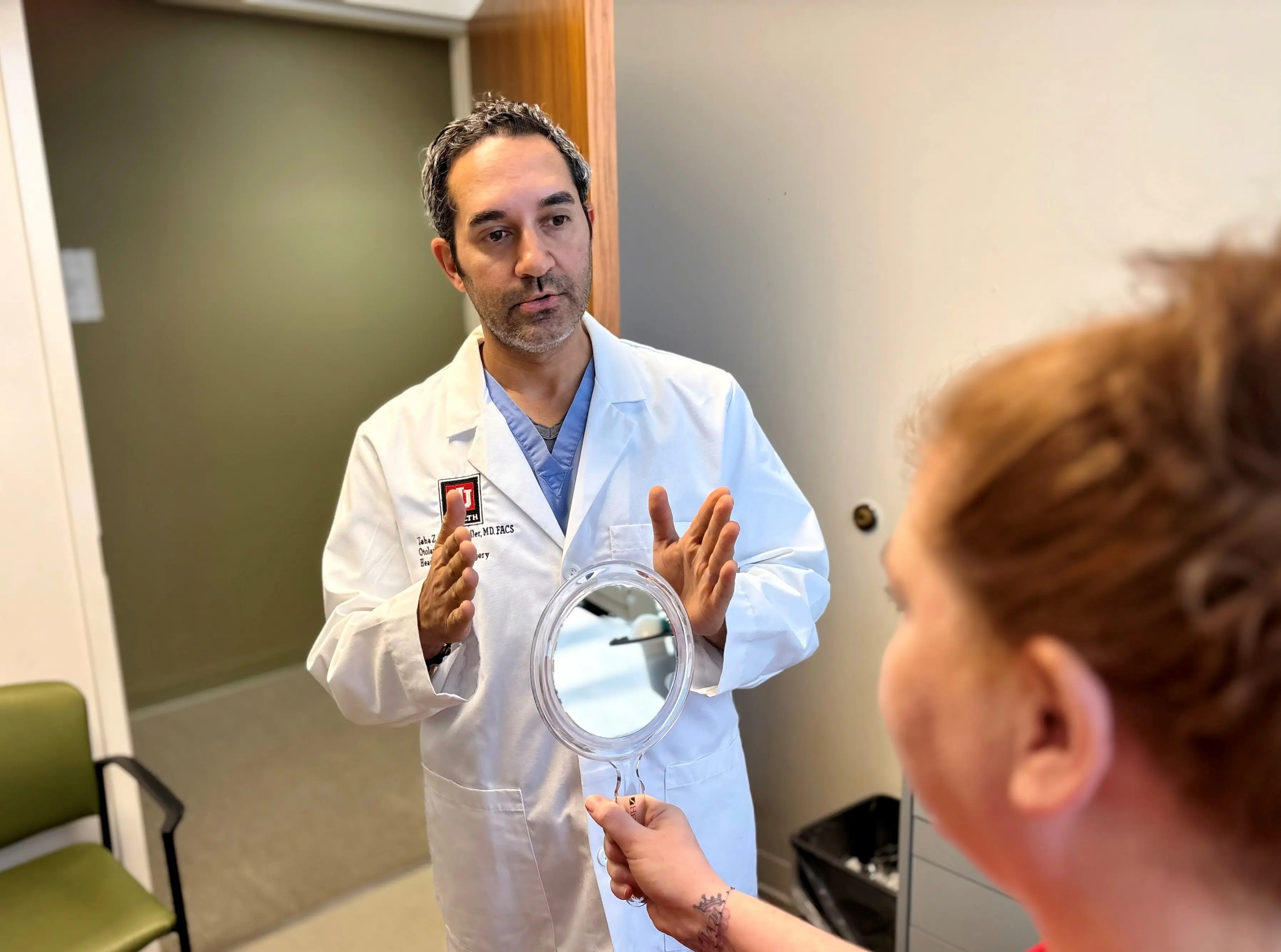 Healthcare provider in a white coat speaks with a patient in an exam room while the patient holds a handheld mirror during a consultation.