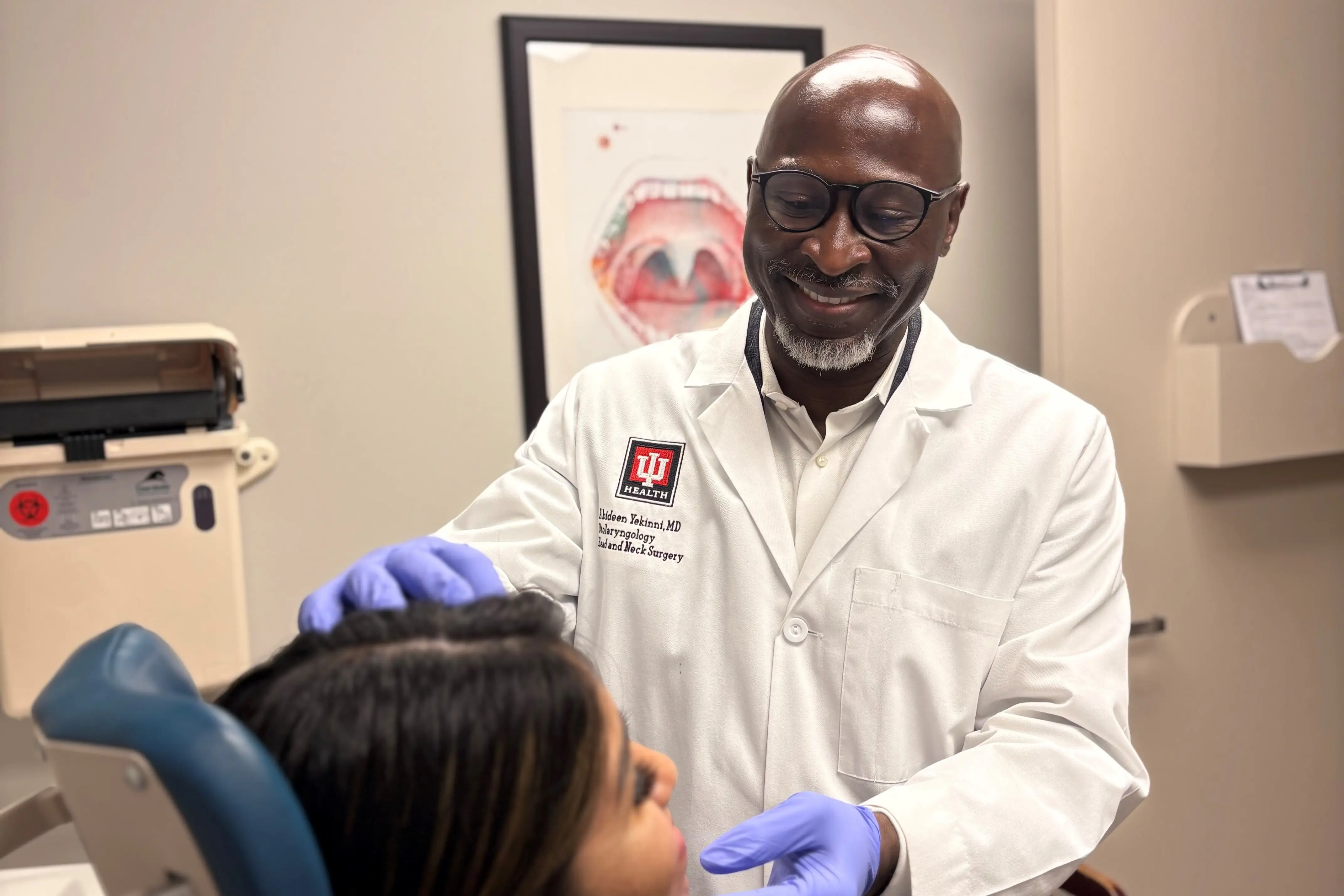 Healthcare provider in a white coat and gloves examines a seated patient in a clinic exam room.