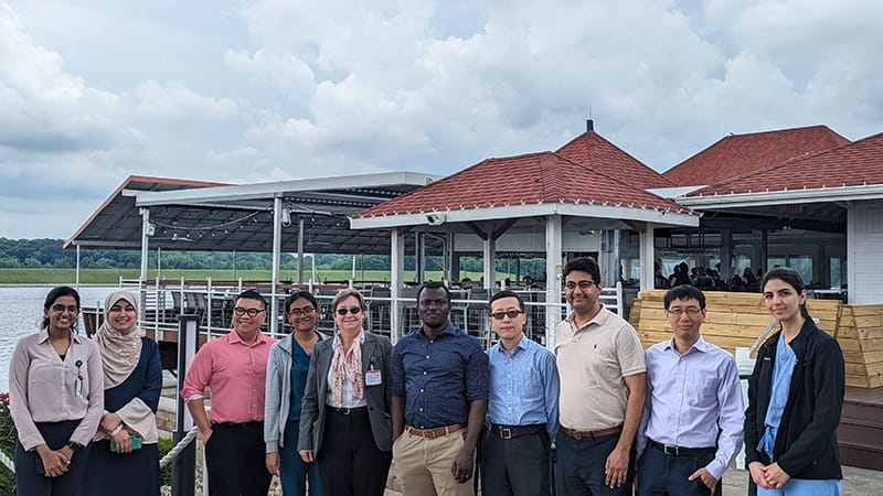 a group of hemepath physicians stand outside on a cloudy day in front of a body of water and a restaurant patio on the dock.