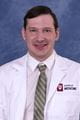 Justin wearing a lab coat and tie. He has short dark hair.
