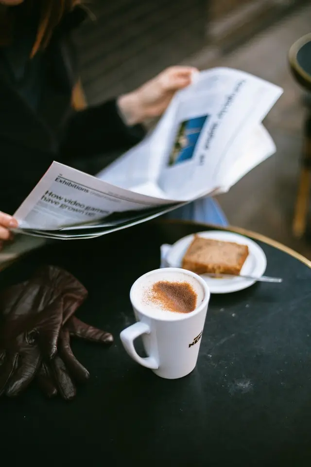 A person at a cafe reads the newspaper. There is a coffee and pastry on the table in front of them.