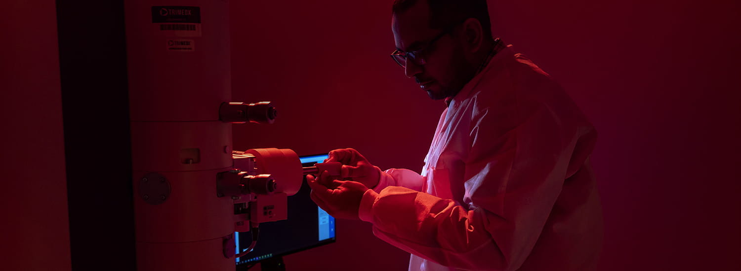 Scientist in a lab coat adjusting a sample on an electron microscope in a red-lit laboratory, with a computer monitor and control panel visible in the background.