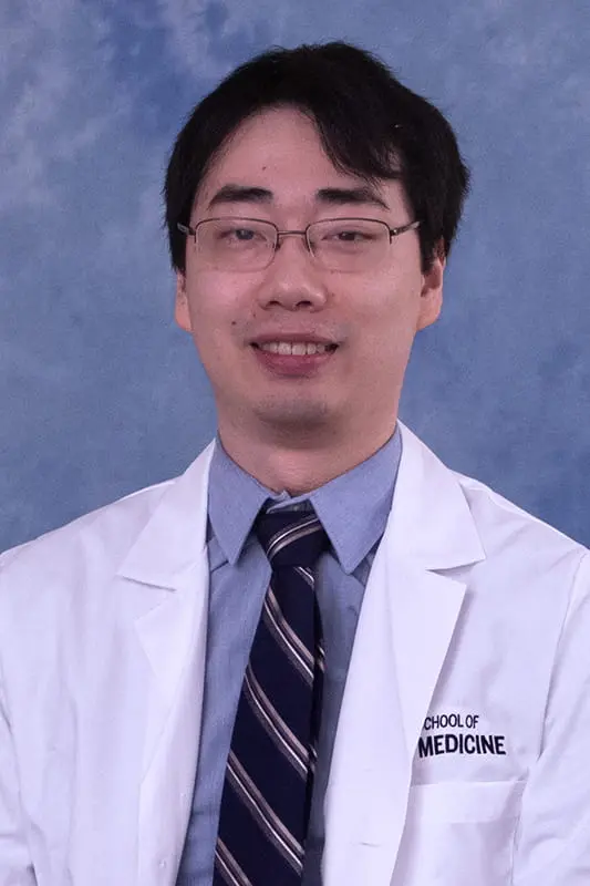Cyrus wearing a lab coat and tie. He has dark short hair and glasses.