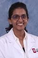 Harini wearing a lab coat over white shirt. She has dark hair pulled back in a ponytail and glasses.