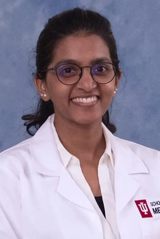 Harini wearing a lab coat over white shirt. She has dark hair pulled back in a ponytail and glasses.