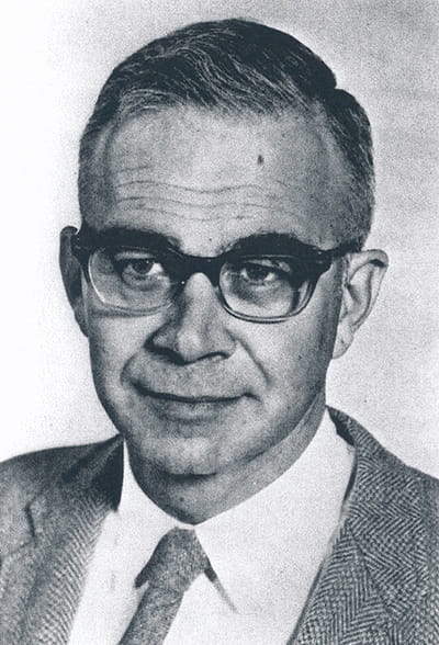 Joshua Edward wears a suit, tie and thick glasses in a black and white portrait