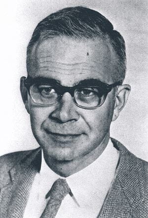 Joshua Edward wears a suit, tie and thick glasses in a black and white portrait