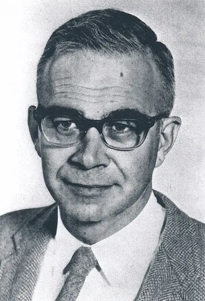 Joshua Edward wears a suit, tie and thick glasses in a black and white portrait