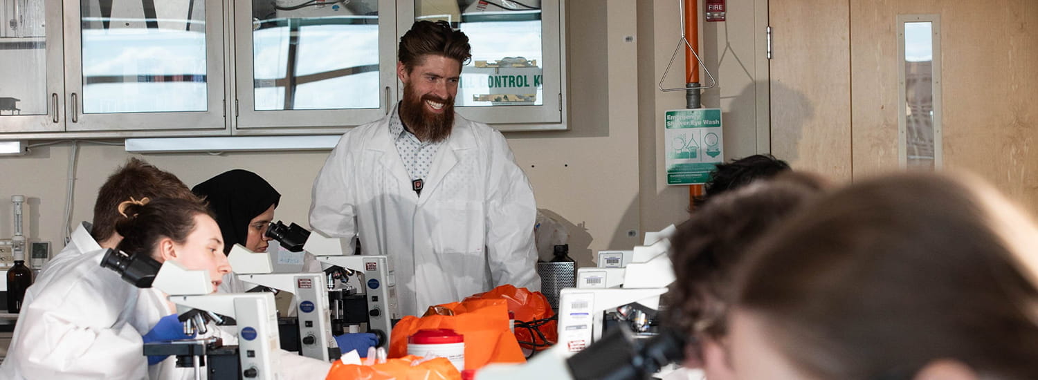A lab instructor, smiling and engaging with students, stands amidst a group of focused individuals using microscopes. The atmosphere in the lab is dynamic, with students intently observing specimens while the instructor provides guidance, fostering an interactive learning environment.