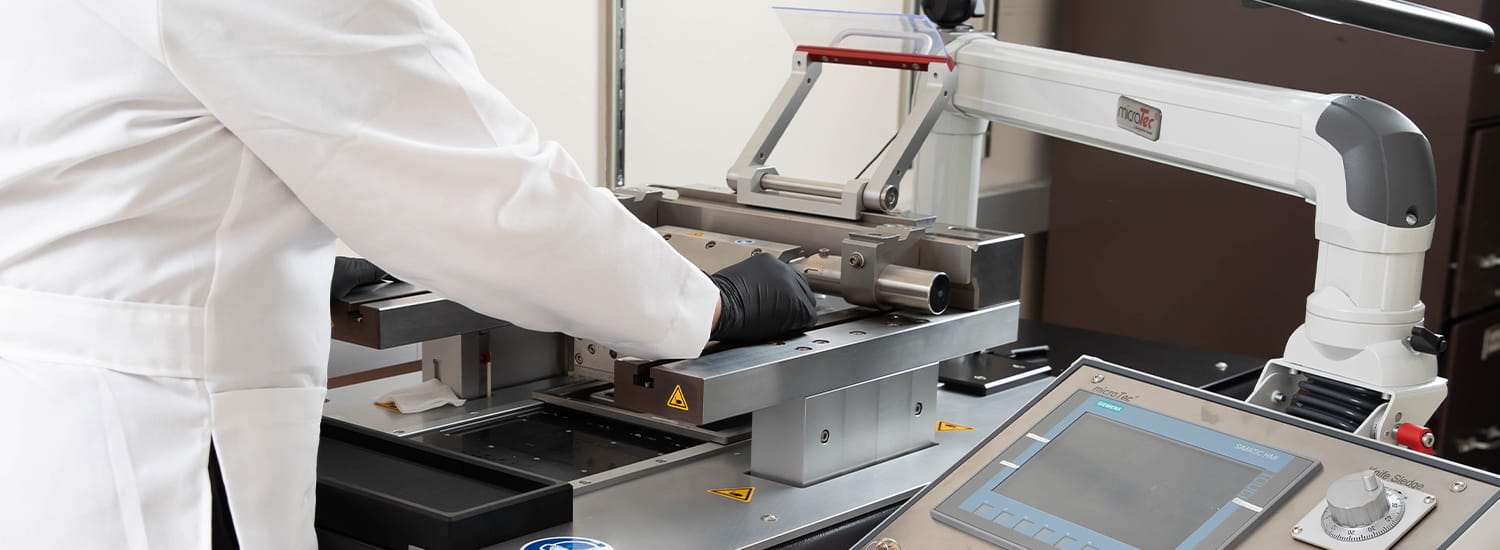 A researcher in a lab coat and black gloves operating a precision laboratory instrument, with a touchscreen control panel and detailed metal components. The equipment appears to be designed for anatomical pathology or tissue processing in a scientific setting.