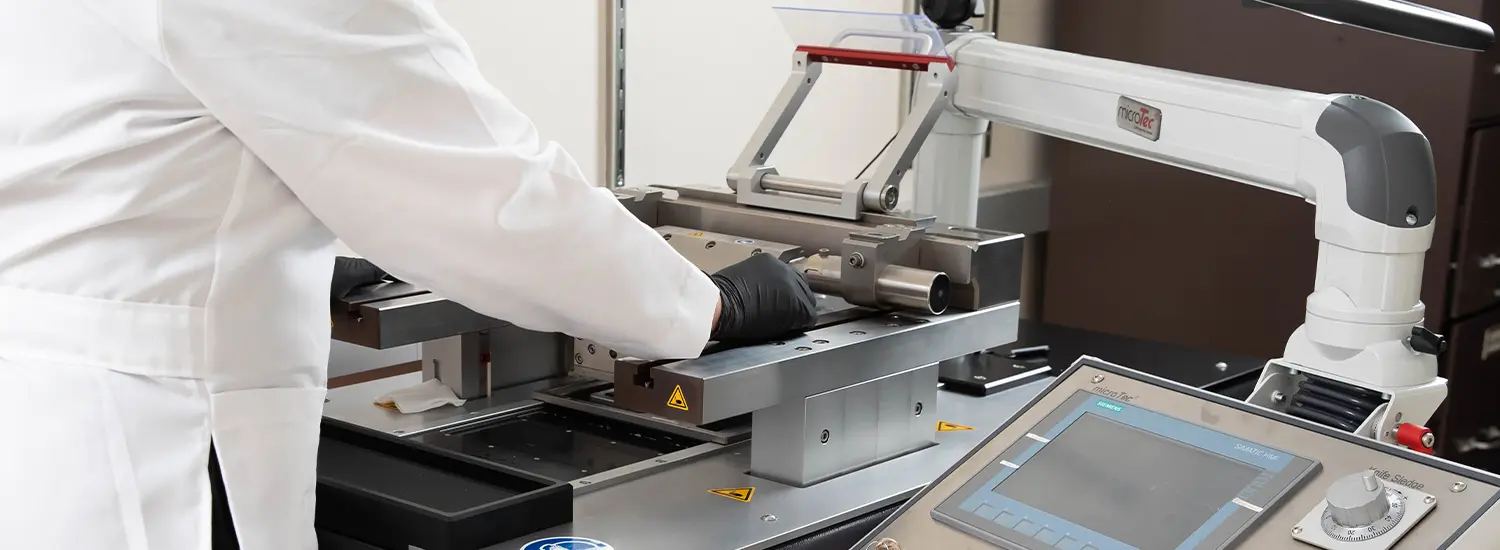 A researcher in a lab coat and black gloves operating a precision laboratory instrument, with a touchscreen control panel and detailed metal components. The equipment appears to be designed for anatomical pathology or tissue processing in a scientific setting.
