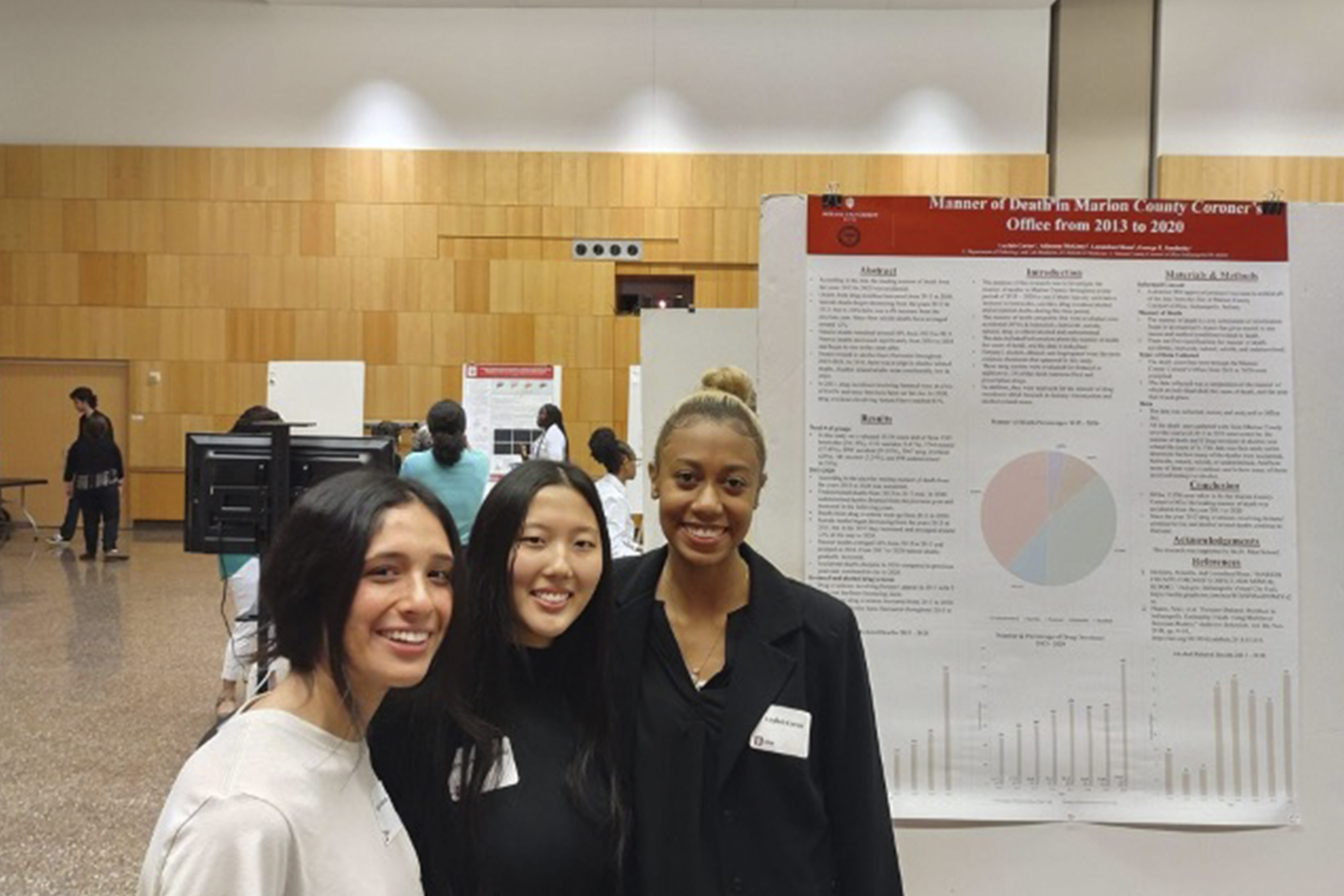 Three young women smiling in front of a research poster presentation in a conference hall. The poster, titled 'Manner of Death in Marion County Coroner's Office from 2013 to 2020,' features graphs and research findings.