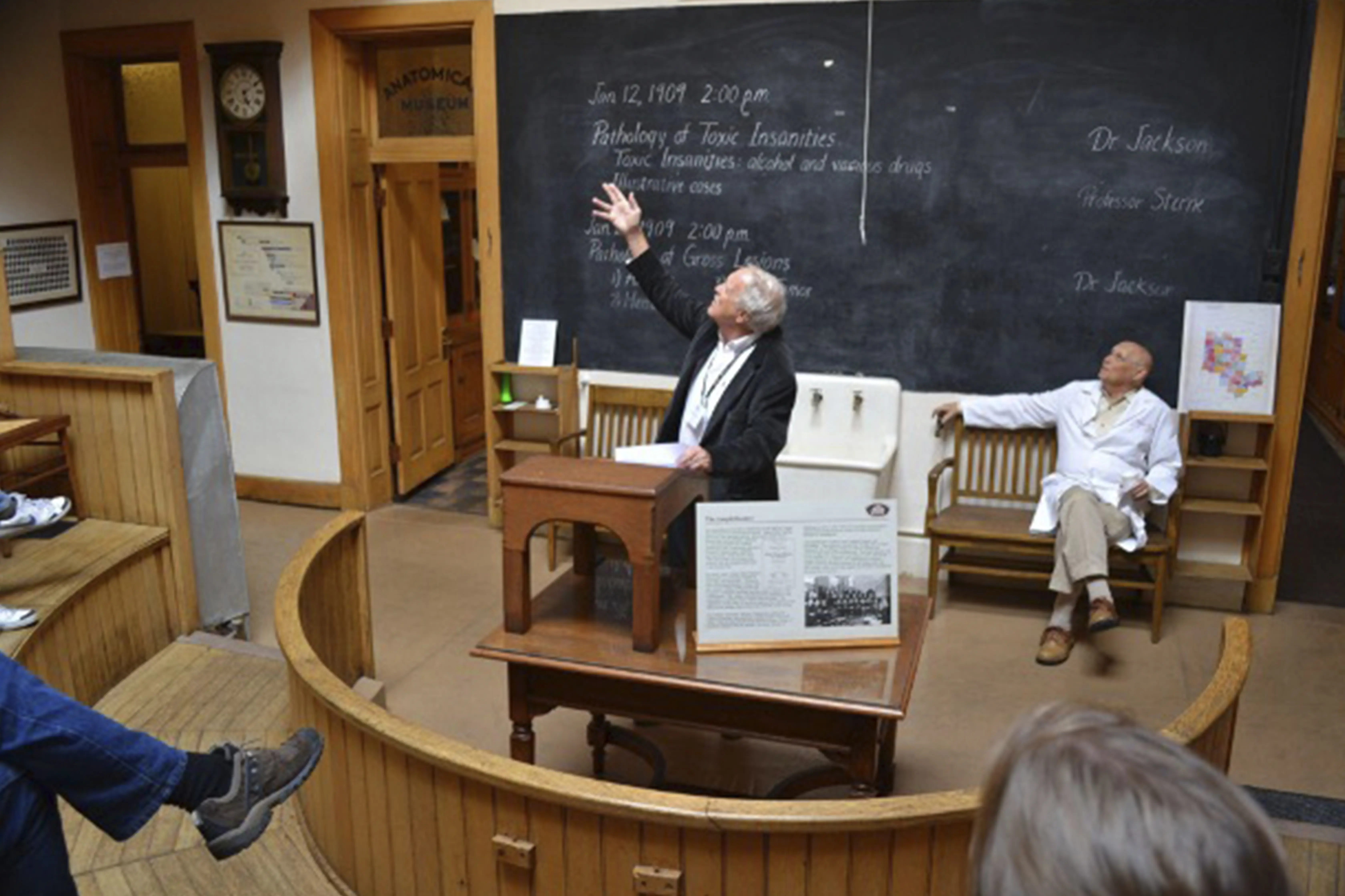 A professor or lecturer gesturing towards a blackboard in a historic anatomical museum or lecture hall. The chalkboard displays an old-fashioned lecture schedule, and another person in a white lab coat sits to the side, observing. 